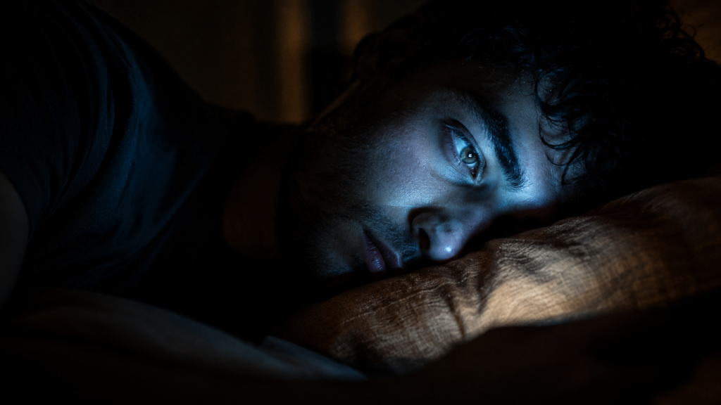 Fotografia cinematográfica em close-up de perfil de uma pessoa deitada no travesseiro em um quarto escuro. Seus olhos estão abertos e estáticos, iluminados apenas pelo brilho azul e frio de uma tela invisível de celular, destacando a expressão de cansaço mental e falta de sono causada pelo uso de tecnologia à noite.